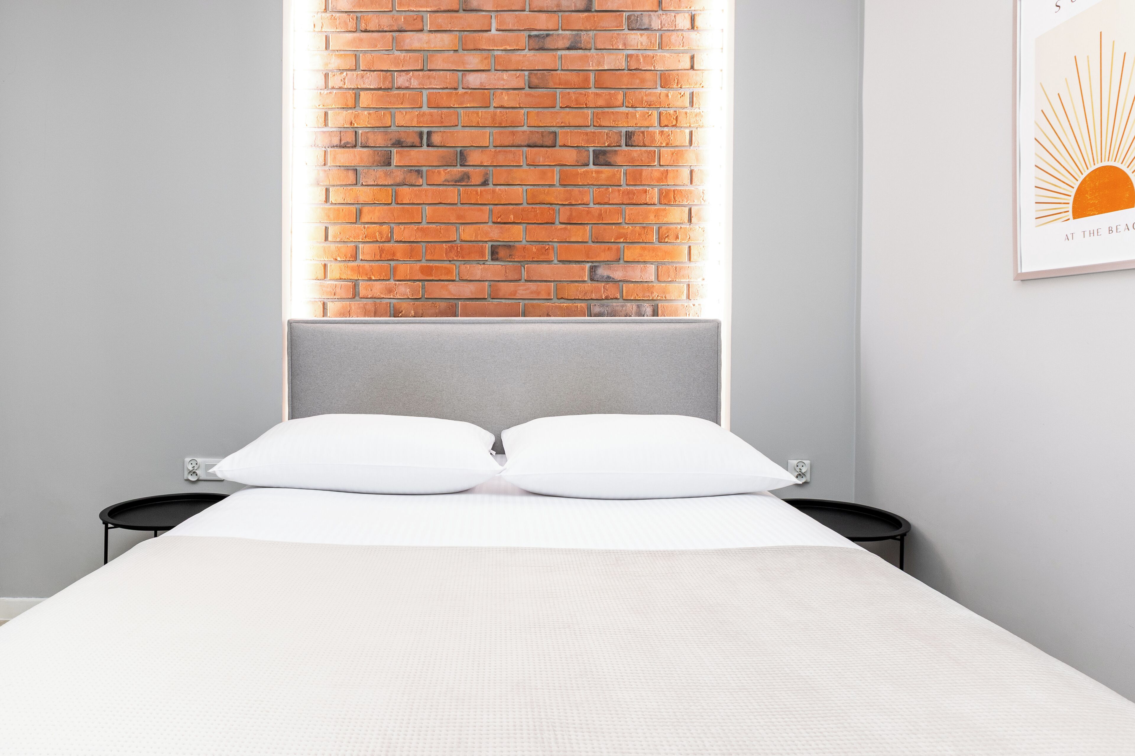 Quarto com parede de tijolos, cama com roupa de cama branca e uma pequena mesa de cabeceira preta.