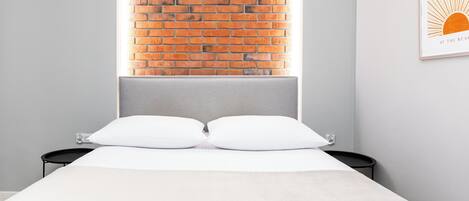 Quarto com parede de tijolos, cama com roupa de cama branca e uma pequena mesa de cabeceira preta.