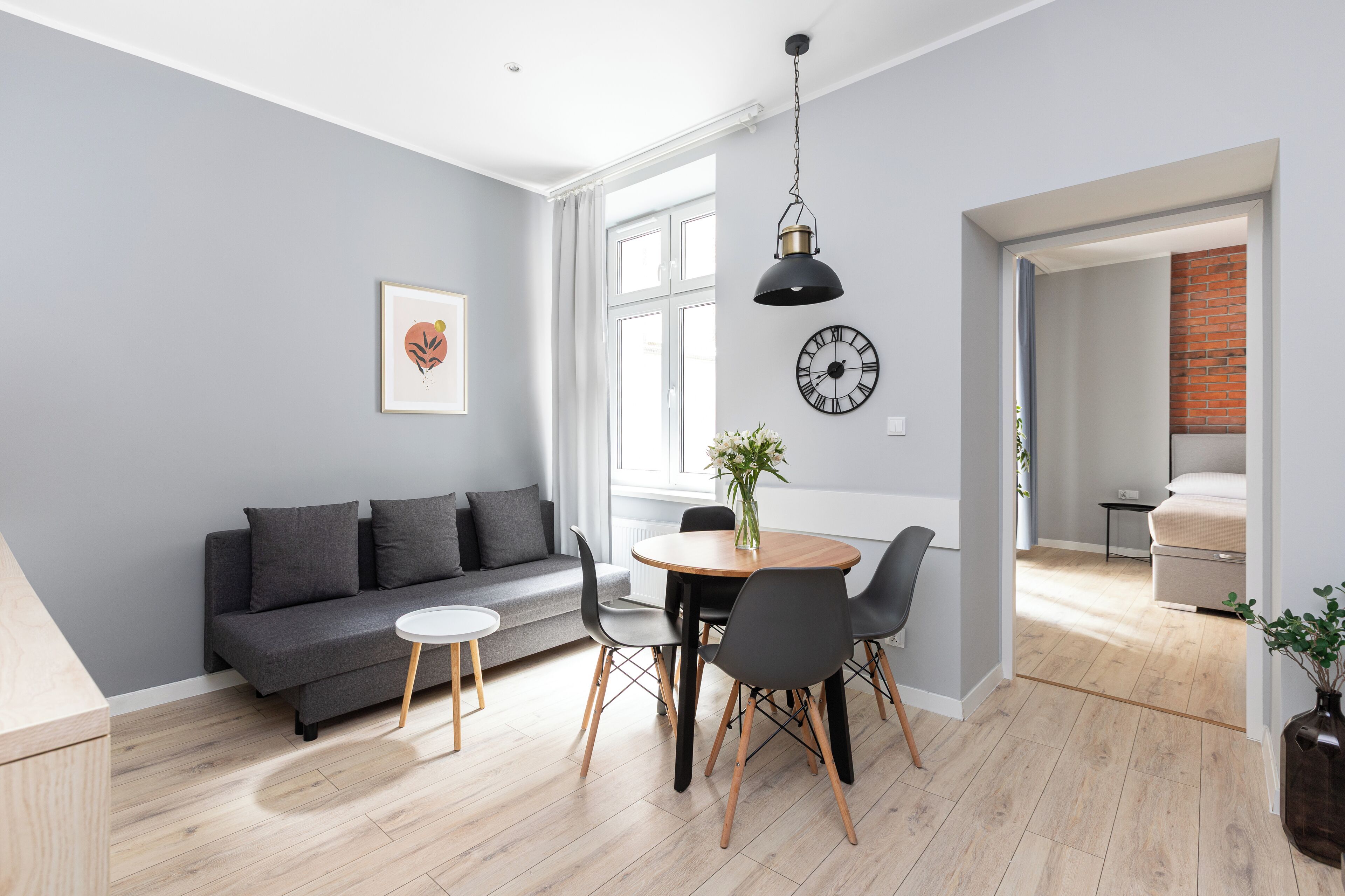 A modern living room with a grey sofa, a round dining table with chairs, a hanging pendant light, and a wall clock.