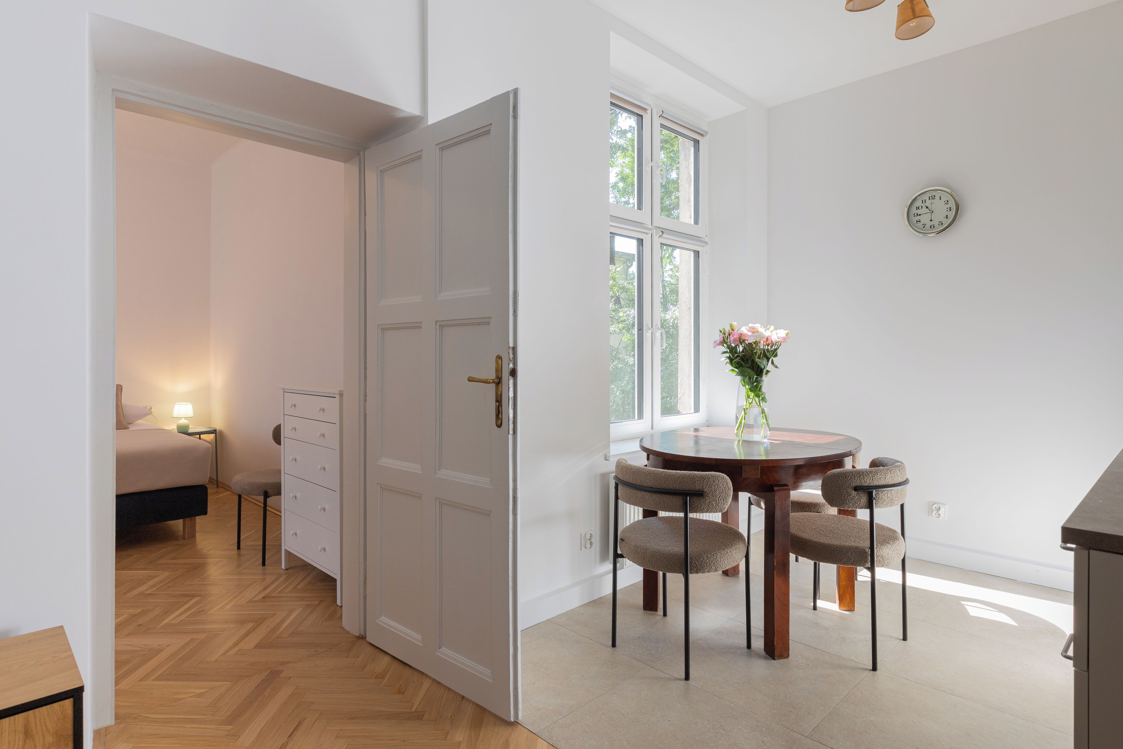 Una habitación con suelo de parqué, una puerta blanca, una mesa de comedor con sillas y un reloj en la pared.