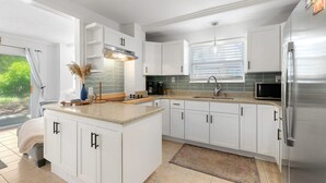 A modern kitchen with white cabinets, a stainless steel refrigerator, and a window with blinds. - Turtle Hideaway- Walk to Beach & Pier! (St. Augustine)