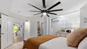 A bedroom with a bed, a ceiling fan, a window with blinds, a bedside table with a lamp, and a door leading to another room. - Turtle Hideaway- Walk to Beach & Pier! (St. Augustine)