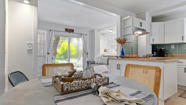 A modern kitchen with white cabinets, a dining area with a round table, and a view of greenery outside. - Turtle Hideaway- Walk to Beach & Pier! (St. Augustine)