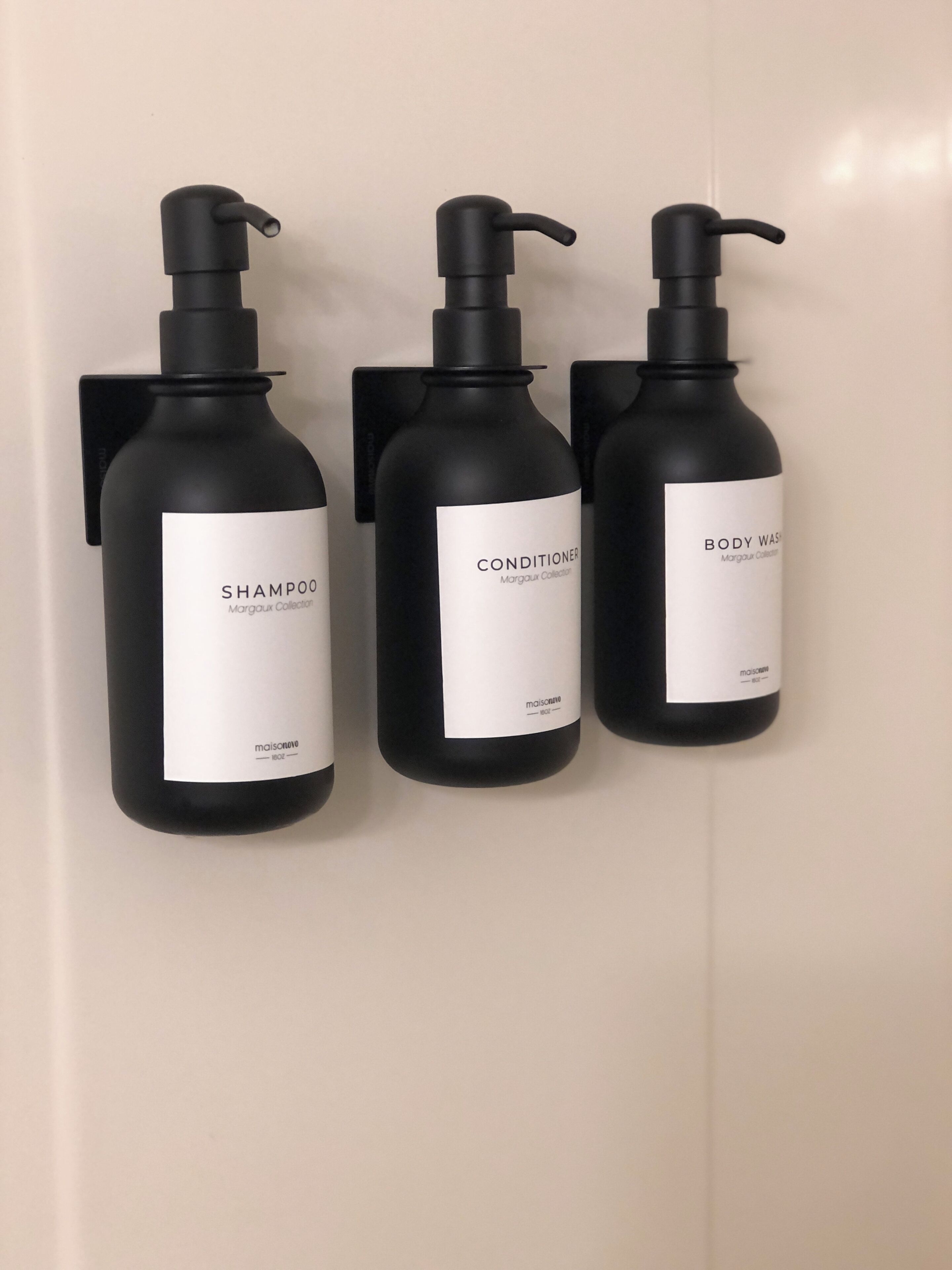 Drie zwarte flessen shampoo, conditioner en douchegel met pompdispensers, gelabeld 'MARGOUX COLLECTION', bevestigd aan een muur.