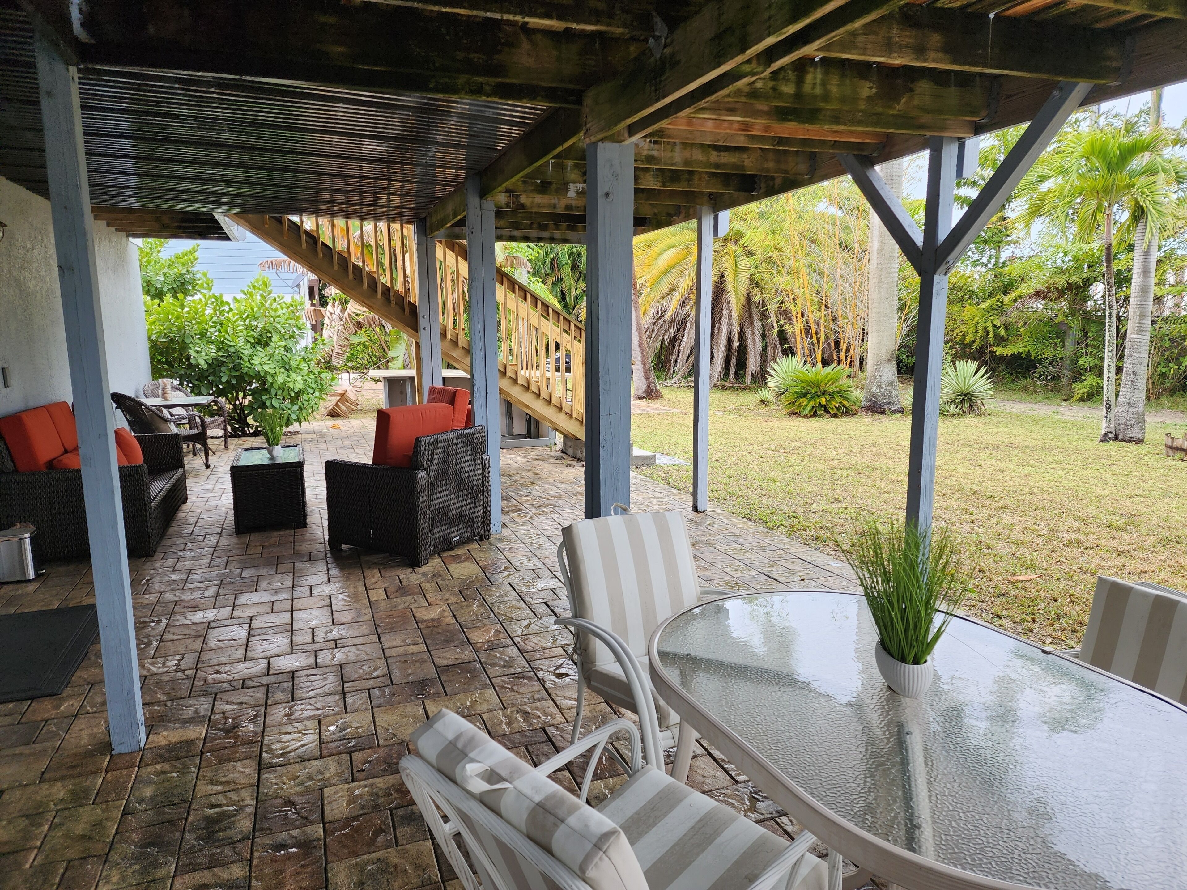 A covered patio area with wicker furniture, a glass table, and a view of a garden with palm trees.