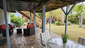 Un patio couvert aménagé avec des meubles en osier, une table en verre et une vue sur un jardin agrémenté de palmiers.