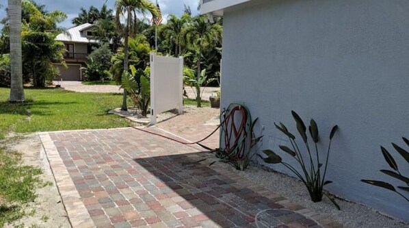 Um caminho de paralelepípedos leva a um edifício com parede externa branca, uma pequena área de jardim com plantas e uma porta de garagem visível.