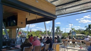 Un espace de restauration en plein air avec des chaises et des tables en métal, un comptoir en bois où est affiché le menu, et une vue dégagée sur des palmiers et un ciel bleu.