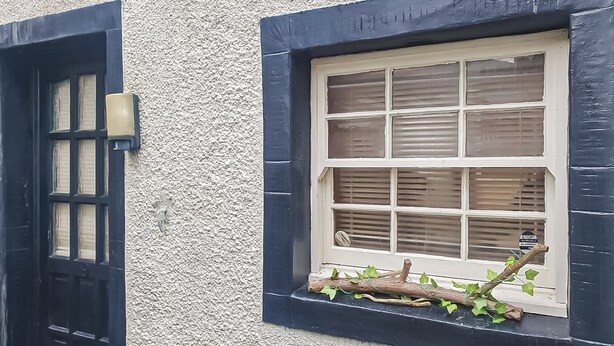 A building facade with a textured wall, a black door with a glass panel, and a window with multiple panes and a branch decoration. - 1 bedroom accommodation in Workington (Workington)