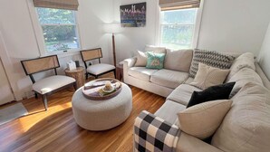 A living room with a beige sofa, a round ottoman, a side table, a chair, and a lamp. There are two windows with curtains.