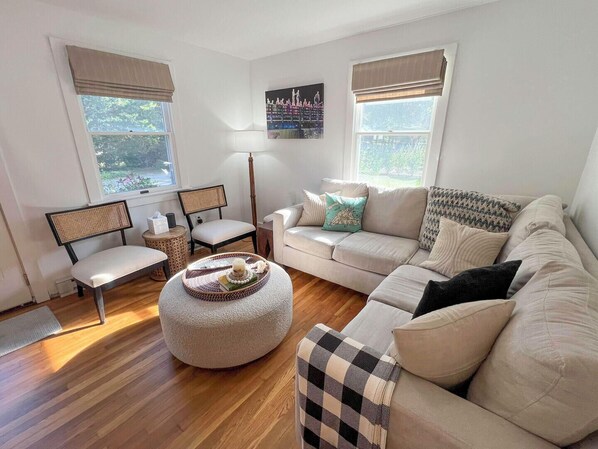 A living room with a beige sofa, a round ottoman, a side table, a chair, and a lamp. There are two windows with curtains. - COZY 3 BR/2 BA CAPE COD IN QUIET NEIGHBORHOOD. CLOSE TO OB CENTER AND LAGOON. (Oak Bluffs)