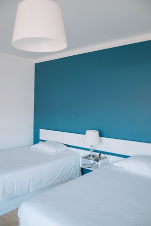 A hotel room with two beds, a white headboard, a bedside table with a lamp, and a large white ceiling light. - ARCA NOVA GUEST HOUSE & HOSTEL (Caminha)