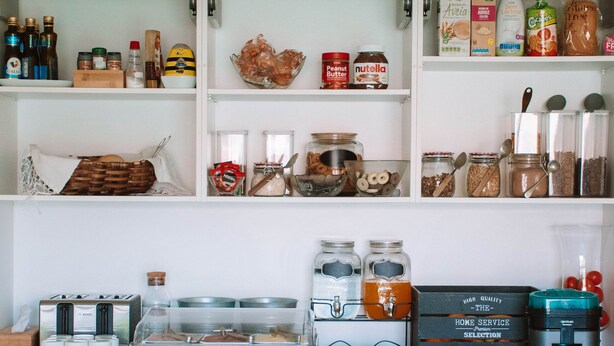 A kitchen with white cabinets, shelves stocked with food items, and a coffee station. - ARCA NOVA GUEST HOUSE & HOSTEL (Caminha)