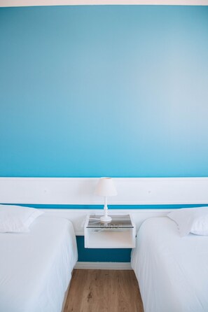 A hotel room with two beds, a white nightstand with a lamp, and a blue accent wall. - ARCA NOVA GUEST HOUSE & HOSTEL (Caminha)