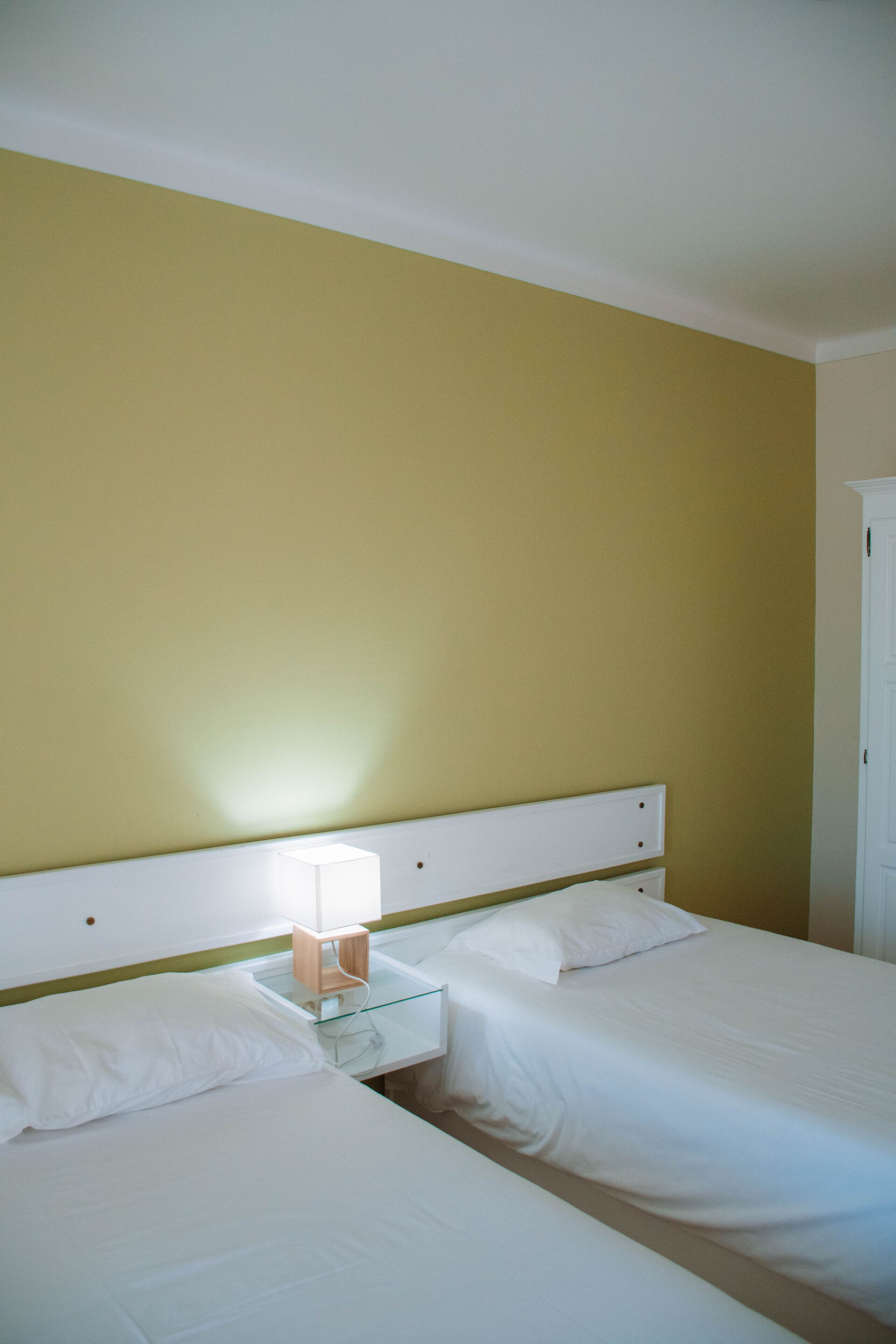 Habitación de hotel con dos camas, una mesita de noche con lámpara y una puerta blanca.
