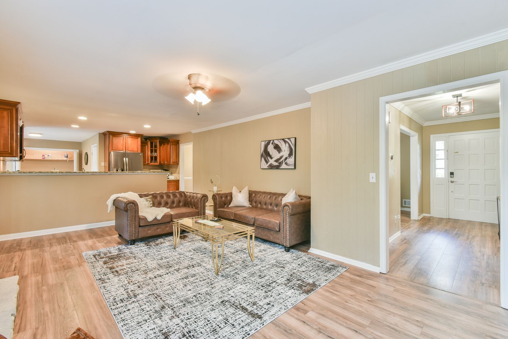 A spacious living room with a large brown leather sofa, a glass coffee table, and a patterned rug. There is a kitchen area with wooden cabinets and a refrigerator in the background.