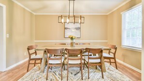 A dining room with a large wooden table, six chairs, a chandelier, and a window with blinds.