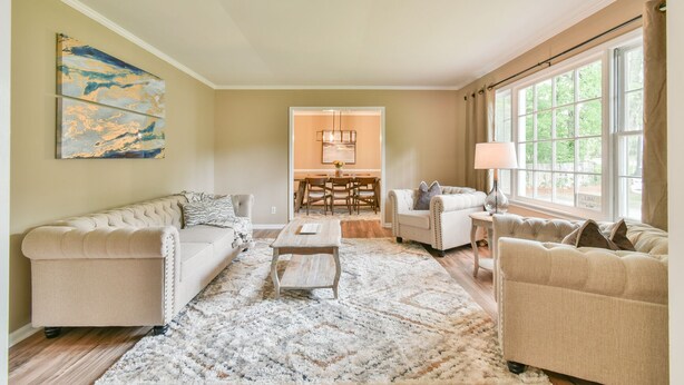 A living room with a sofa, armchairs, a coffee table, and a rug. There is a dining area in the background with a table and chairs.