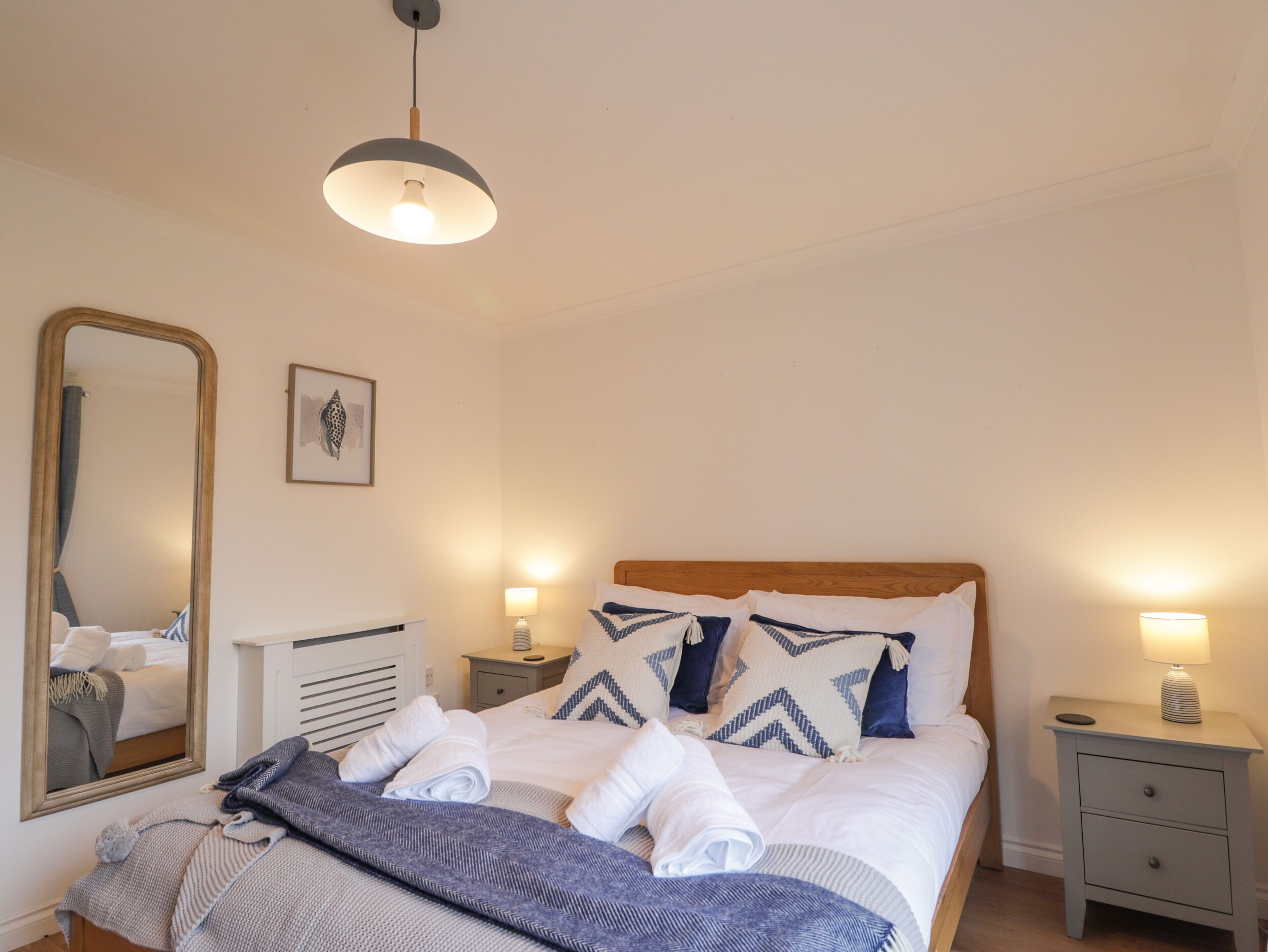 A neatly made bed with pillows, a bedside lamp, a nightstand, a mirror, and a framed picture on the wall.