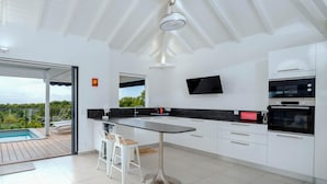 Une cuisine moderne dotée d’un grand îlot central, de meubles blancs et d’un téléviseur à écran plat, avec vue sur l’extérieur.