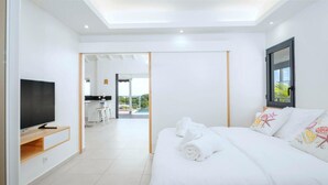 Une chambre d’hôtel moderne avec un grand lit, un téléviseur posé sur un meuble en bois, un balcon donnant sur la piscine et, au fond, une kitchenette.