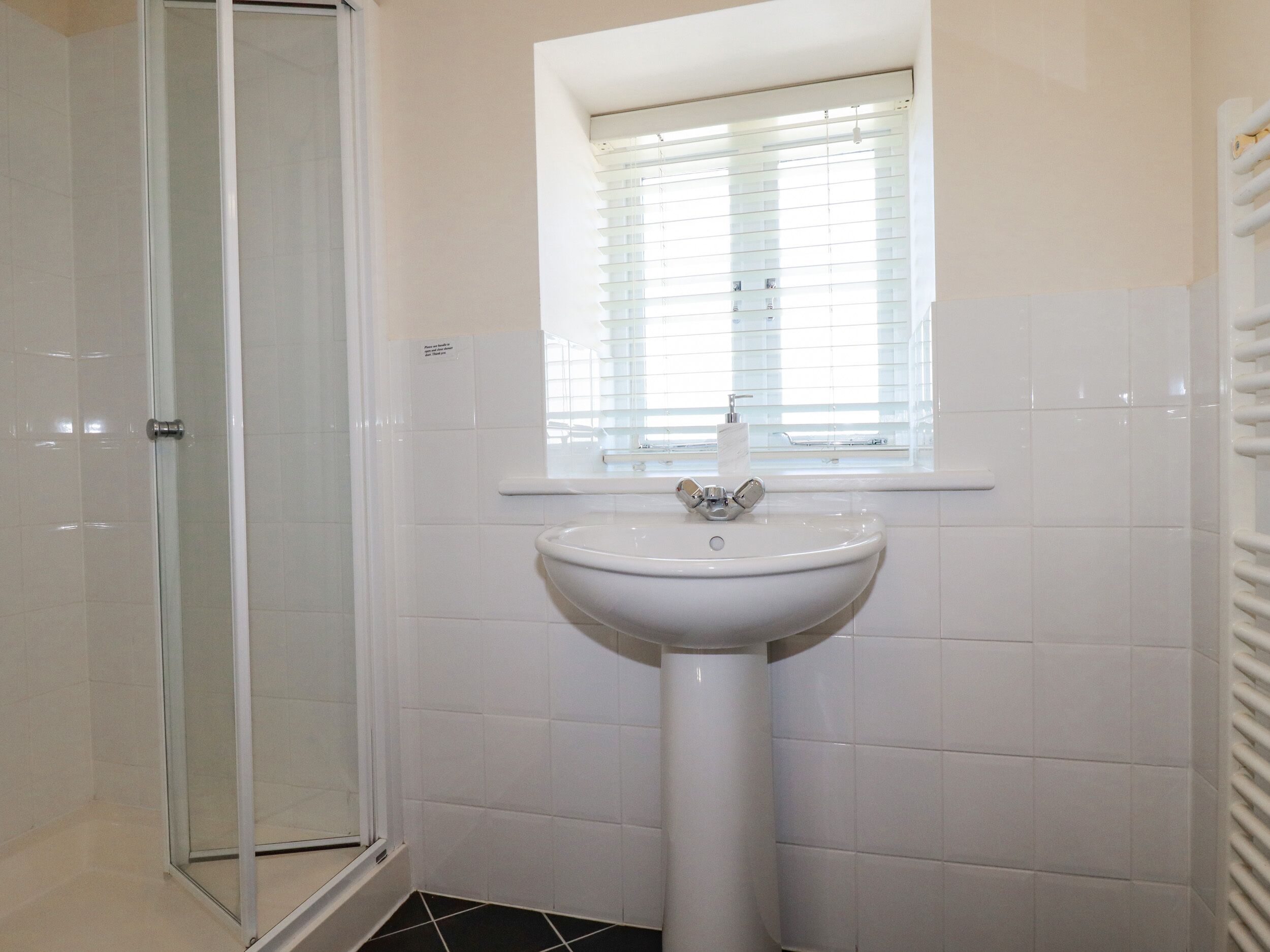 A bathroom with a glass shower enclosure, a white sink, and a window with blinds.