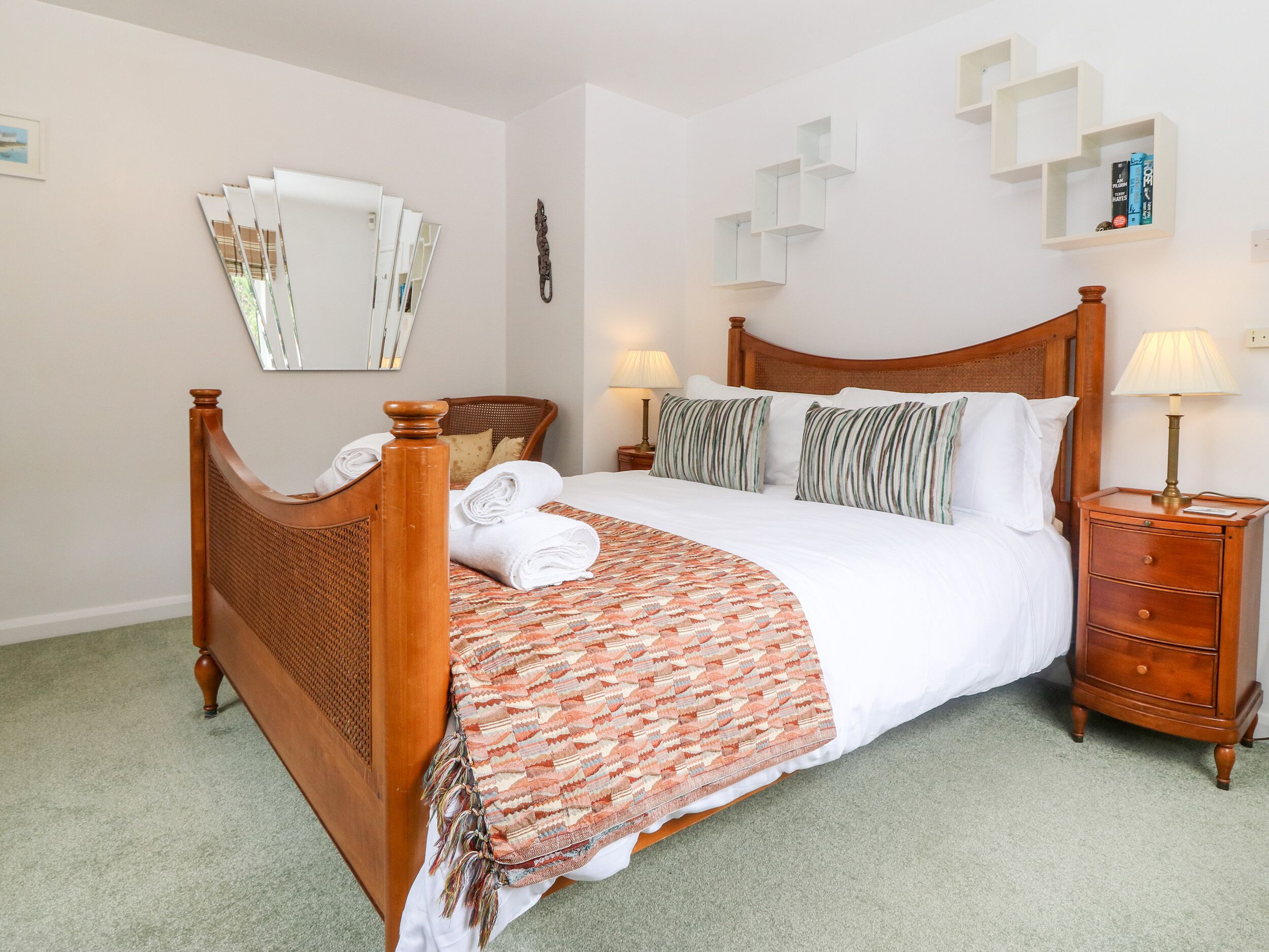 A neatly made bed with a patterned throw, a wooden headboard, a nightstand with a lamp, and a wall-mounted shelf with books.