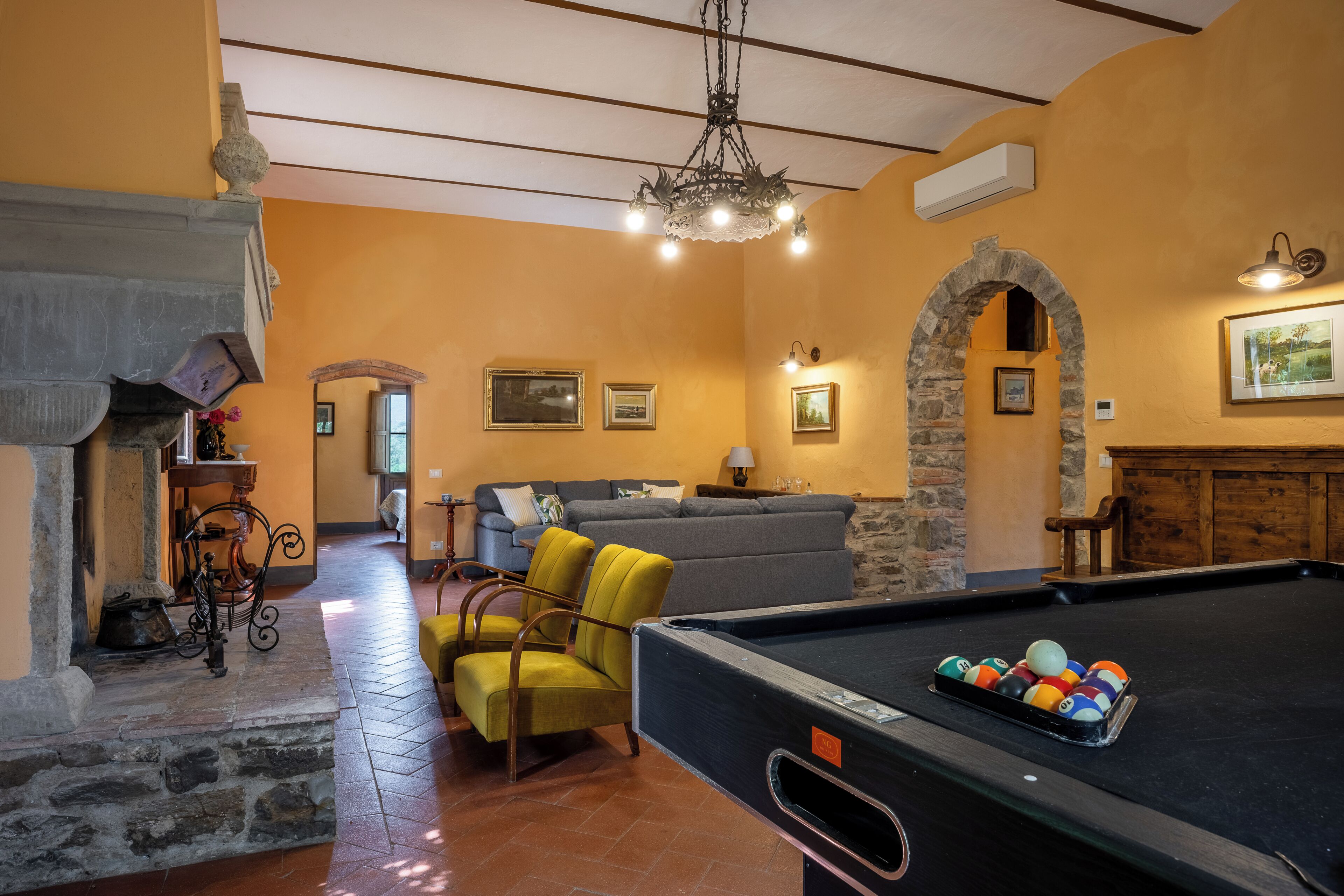 A billiards table, a sofa, a fireplace, and a stone archway in a room with yellow walls and a tiled floor.