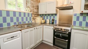 A compact kitchen with white cabinets, a built-in oven, a dishwasher, and a window with a view of greenery. - Myrtle Villa (Knighton)