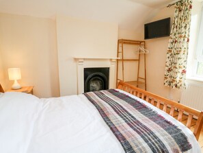 A bedroom with a bed, a bedside table with a lamp, a wooden chair, a fireplace, a TV mounted on the wall, and a window with curtains.