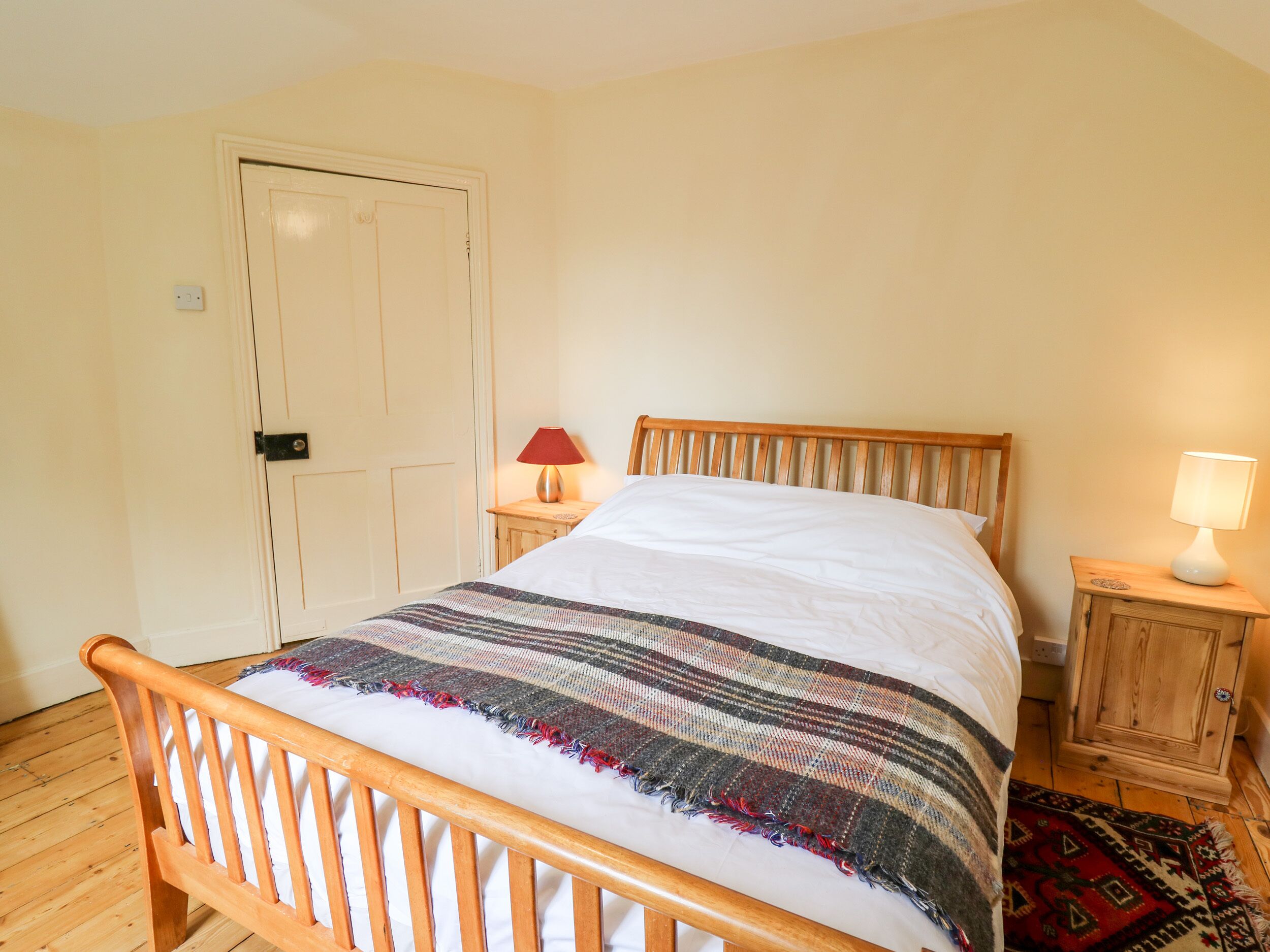 Een slaapkamer met een houten bedframe, een witte matras, een geboeide deken, een rode lamp op een nachtkastje en een houten kast.
