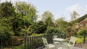 A wooden deck with metal chairs and a table, surrounded by lush greenery and a wooden fence. - Myrtle Villa (Knighton)