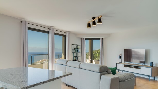 A modern living room with a large grey sofa, a flat-screen TV, and a dining area with a granite countertop. - Banana Tree (Camara de Lobos)