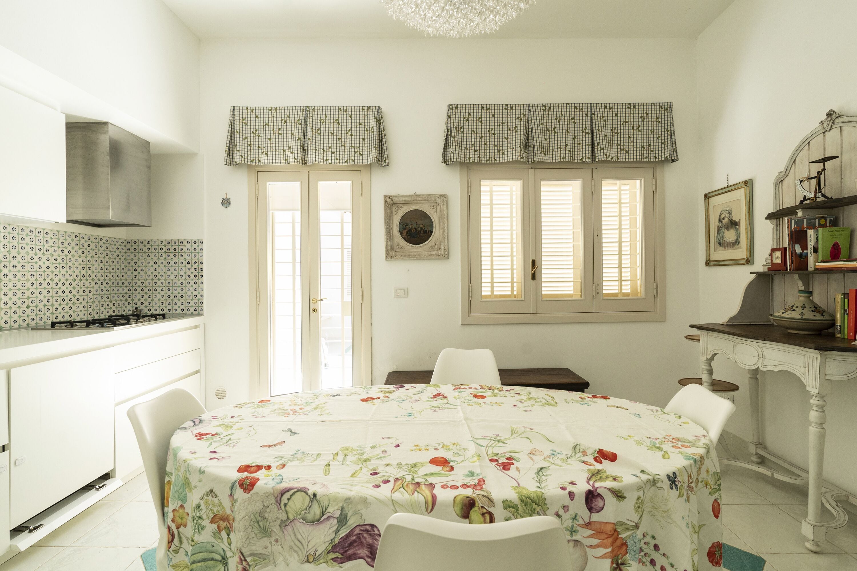Une cuisine avec des meubles blancs, une cuisinière à gaz et une table à manger recouverte d’une nappe à motifs floraux.