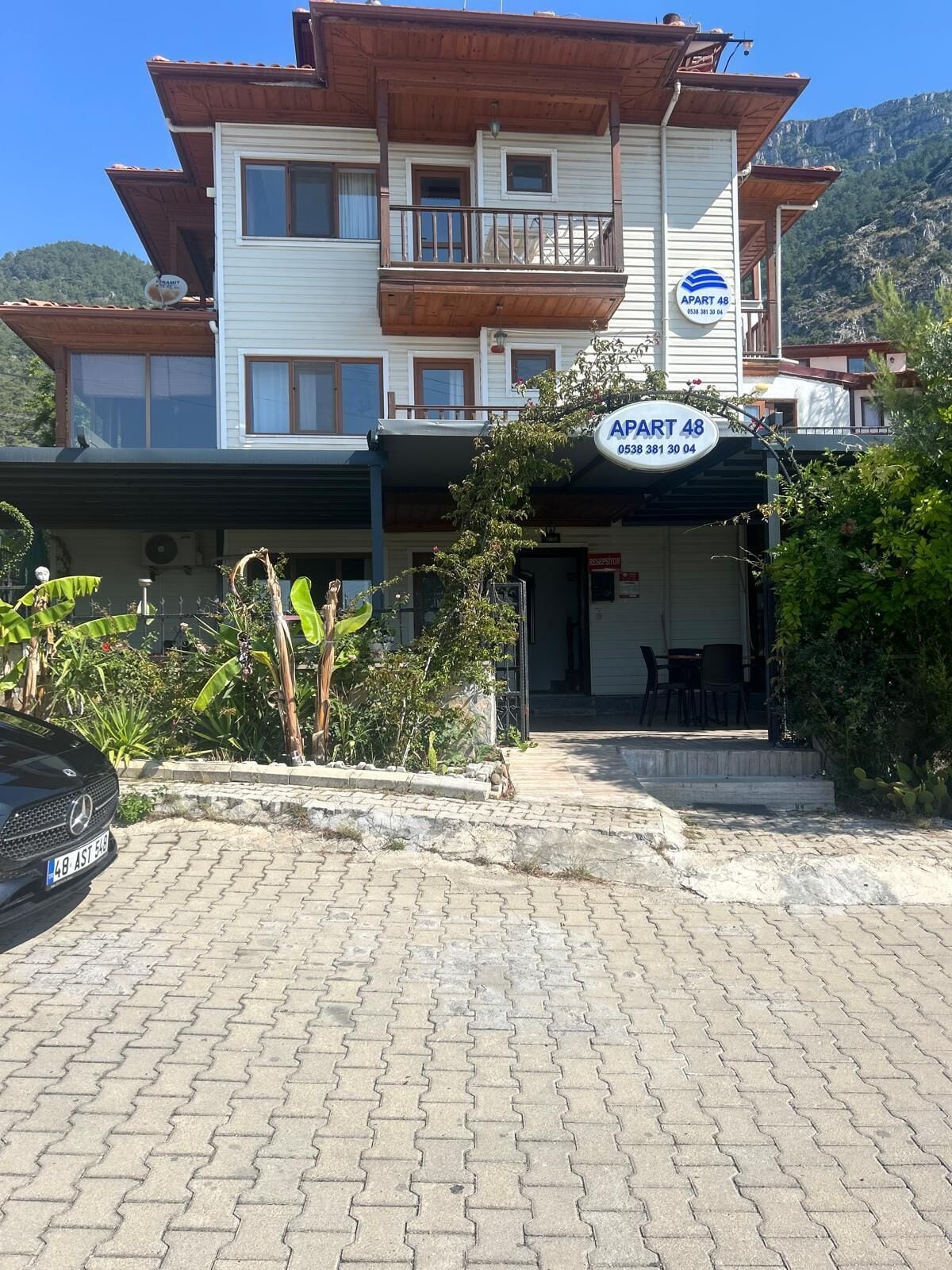 A two-story building with a sign reading "APART 48" and a contact number. There is a paved driveway leading to the entrance, and a car is parked in the foreground.