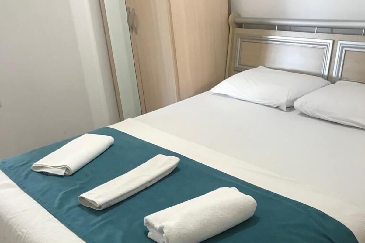 Una cama bien hecha con un edredón verde aguamarina y sábanas blancas, piso de madera y una puerta de madera al fondo.