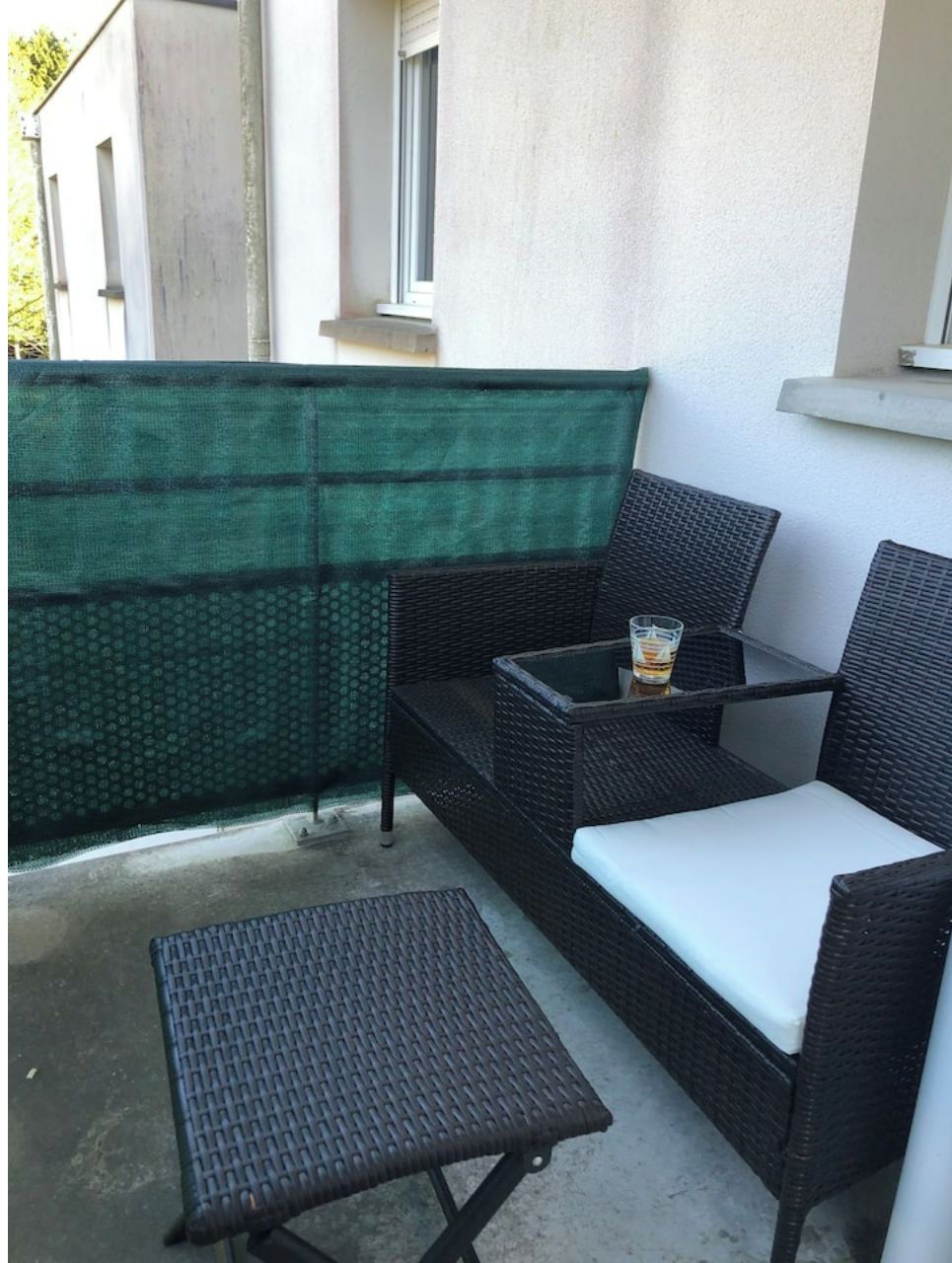 Un balcon agrémenté de meubles en osier, notamment une table et deux chaises, ainsi que d’un paravent en tissu vert.