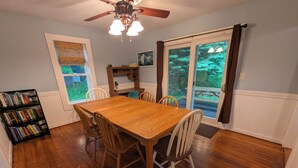 A dining room with a wooden table, chairs, a ceiling fan, a bookshelf, and a window with blinds. - Waldheim Cottage: In-town bar Harbor Hideaway (Bar Harbor)