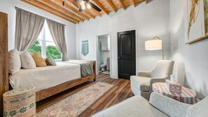 A bedroom with a wooden bed, a sitting area with a chair and a small table, a floor lamp, and a door leading to another room. - Homely Haven Near A&M (Bryan)
