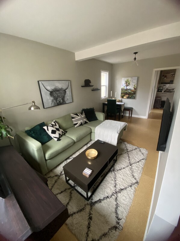 A living room with a green sofa, a coffee table with a bowl and books, a patterned rug, and a dining area with a table and chairs.