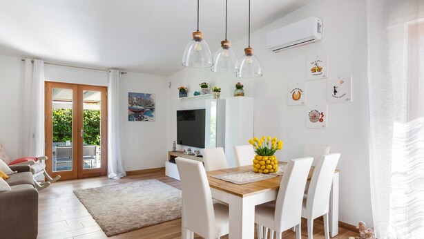 Un salon moderne avec une table à manger en bois, des chaises blanches et un téléviseur. On y trouve un canapé et une table basse sur laquelle est posé un vase de fleurs.