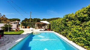 Une piscine rectangulaire bordée de carreaux, entourée d’un chemin de pierre, d’un espace de repos couvert et d’une végétation luxuriante.