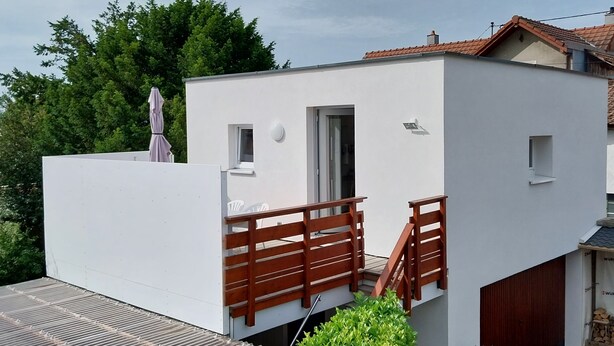 A modern white building with a flat roof, a small balcony with wooden railings, and a door leading to an interior space.