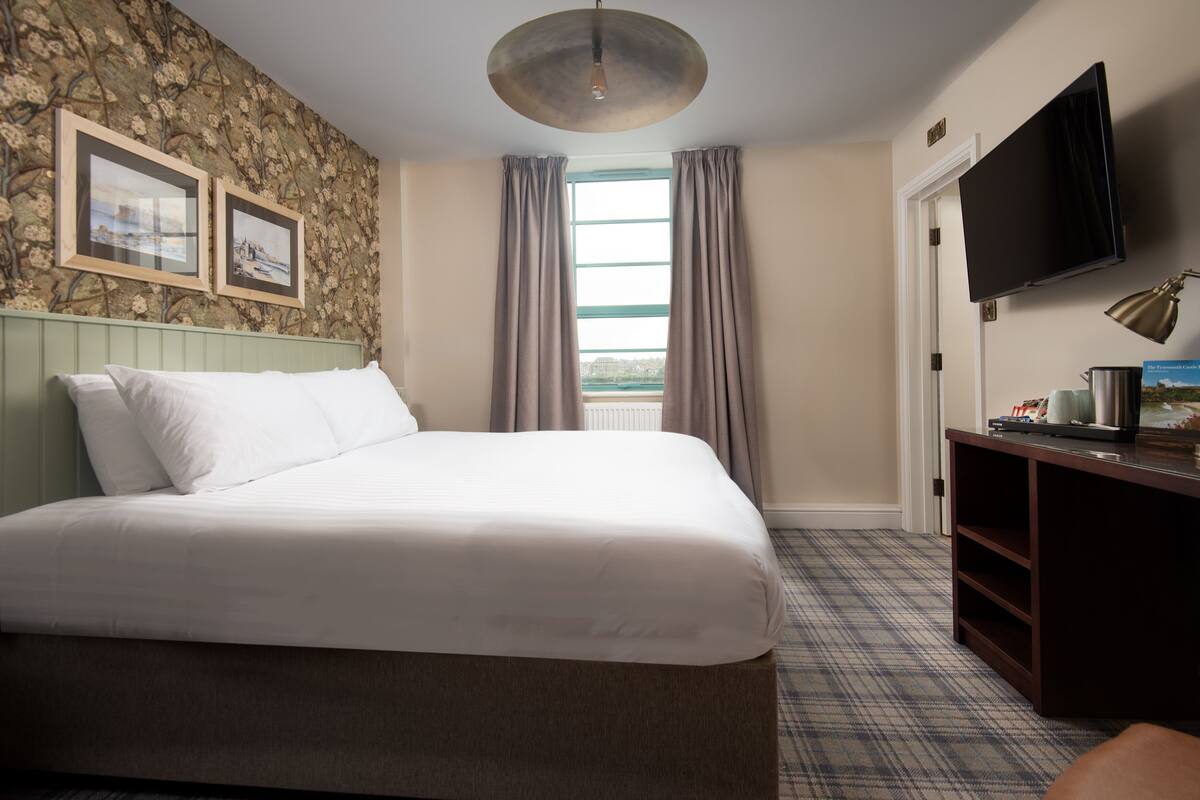 A hotel room with a bed, a television, a desk with a lamp, and a window with curtains.