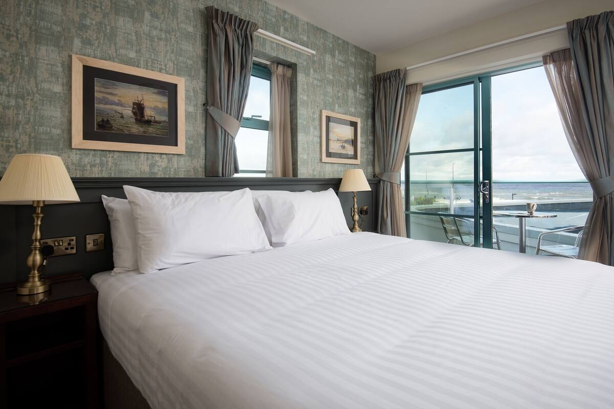 A hotel room with a large bed, two bedside lamps, a framed picture on the wall, and a view of the sea through the window.