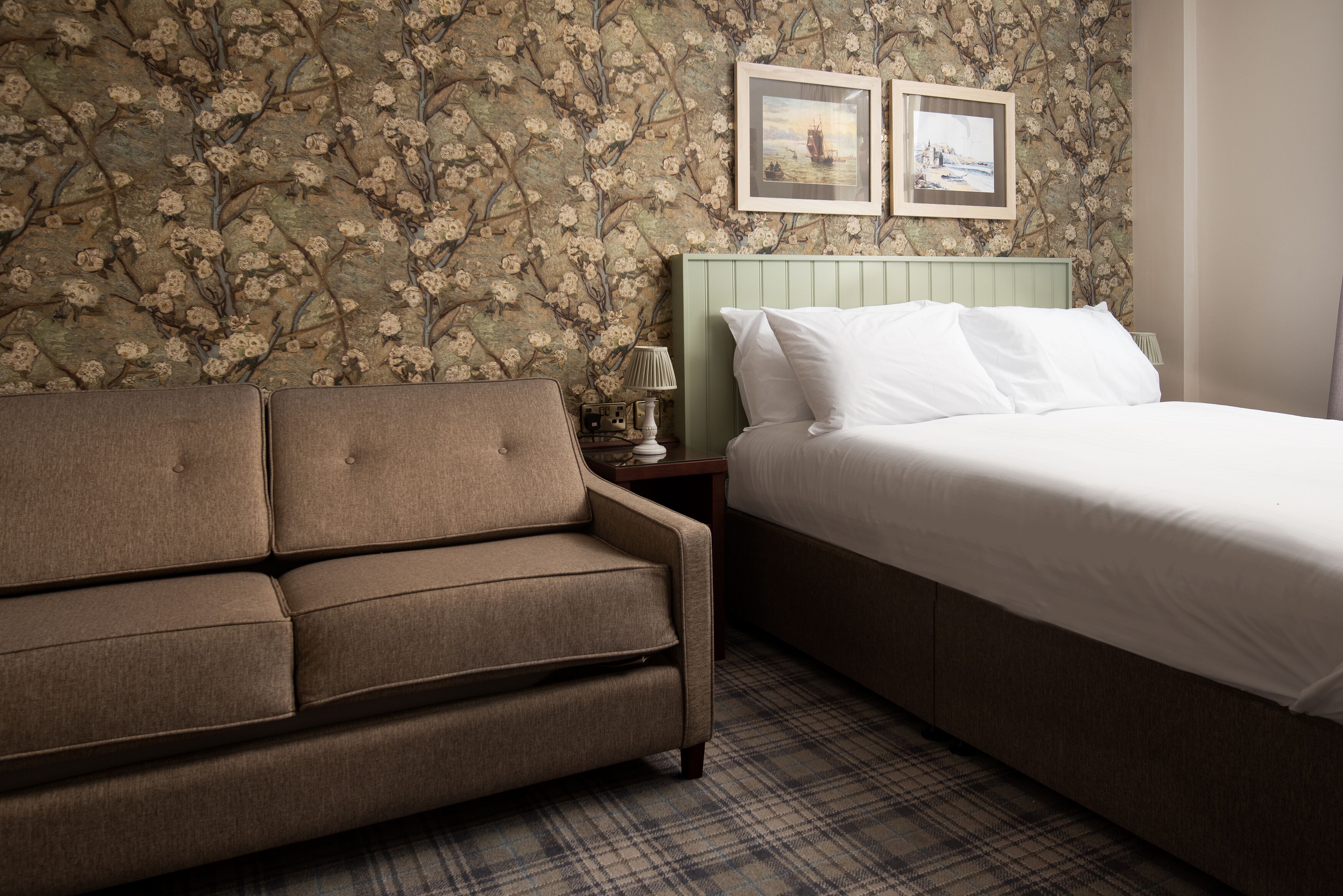 Quarto com papel de parede floral, cabeceira verde, sofá marrom, cama com roupa de cama branca e duas imagens emolduradas na parede.