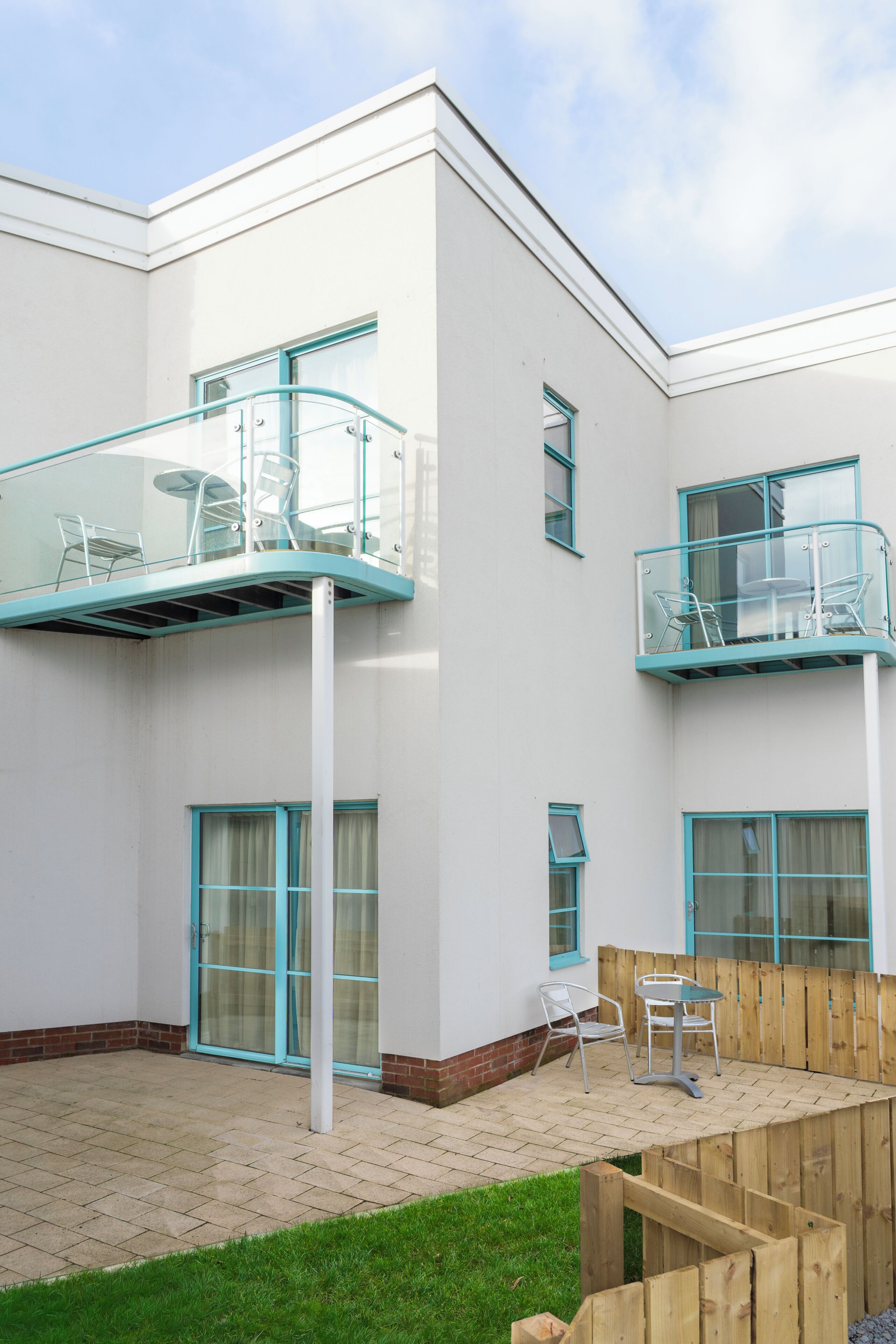Ein zweistöckiges Gebäude mit einem Glasbalkon, einer Terrasse mit Sitzgelegenheiten und einem Holzzaun.