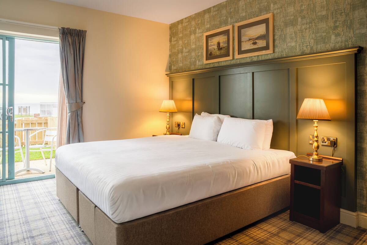 A hotel room with a large bed, two bedside lamps, a view through a glass door, and two framed pictures on the wall.
