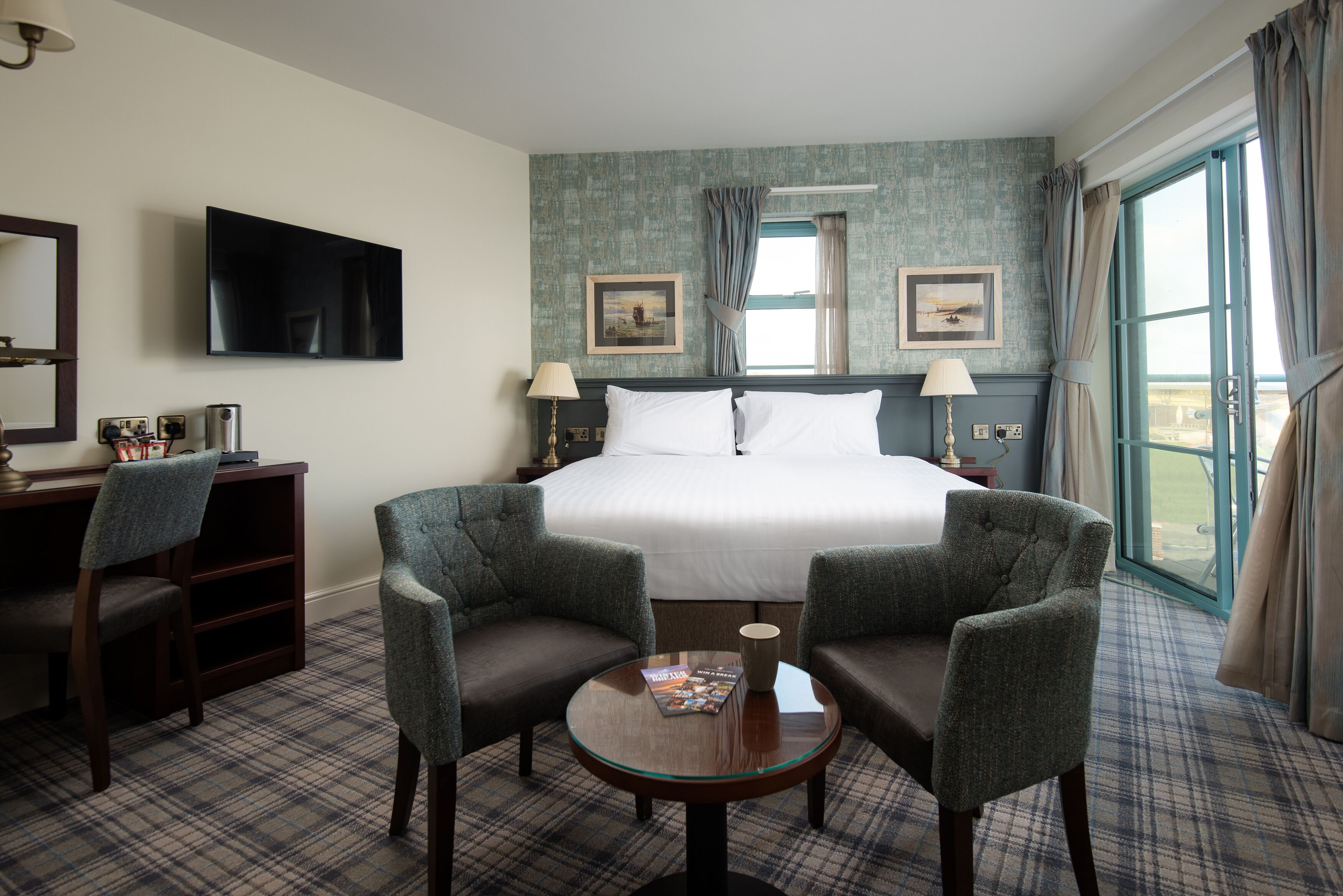 A hotel room with a bed, two armchairs, a small table, a TV, and a window with curtains.