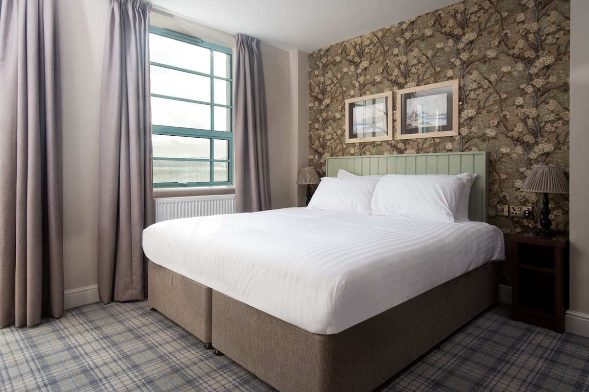 A neatly made bed with a headboard, a window with curtains, and a floral wallpaper.